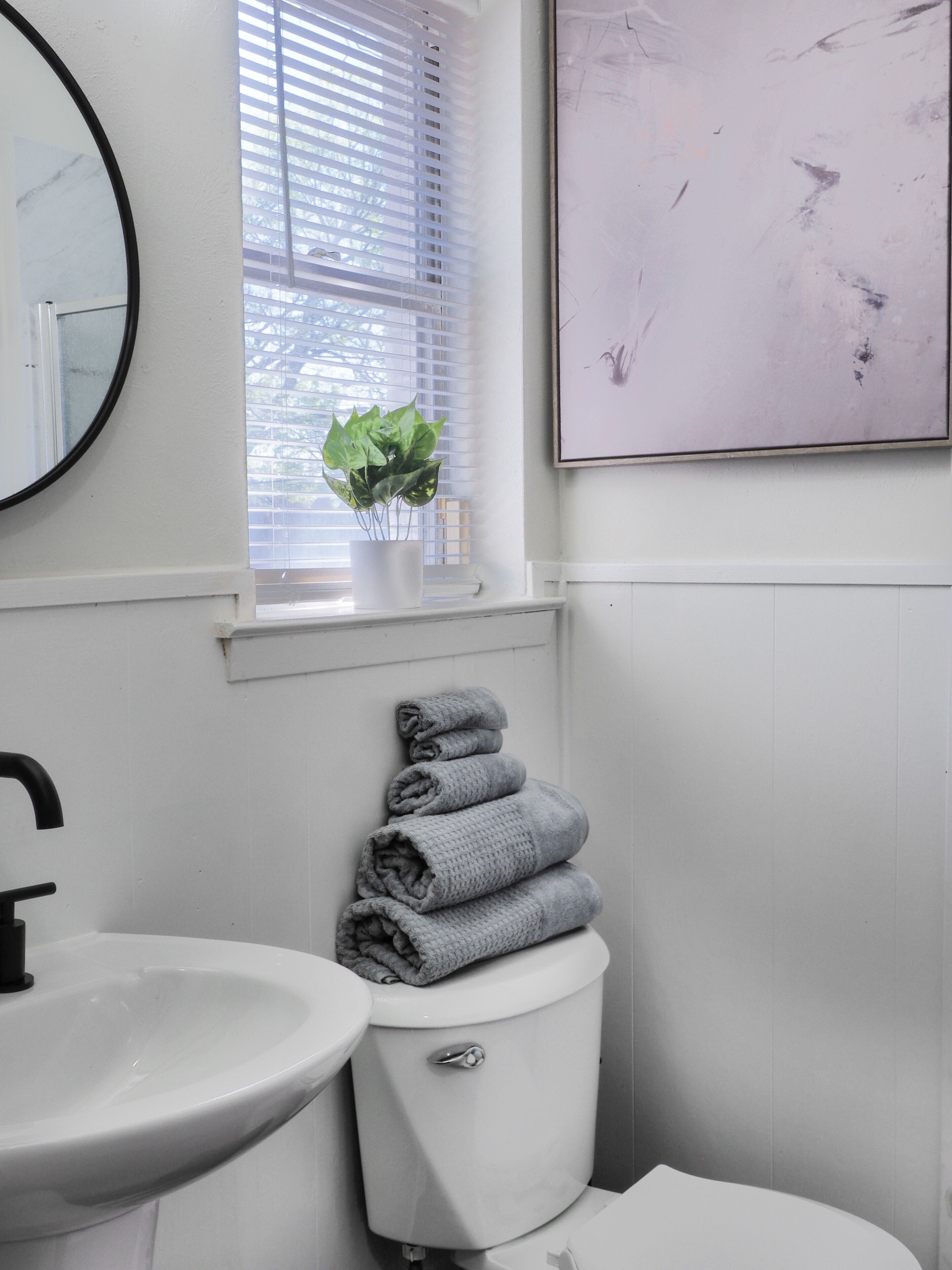 A bathroom with a white sink, a toilet, a window with blinds, a potted plant, and a framed picture on the wall.