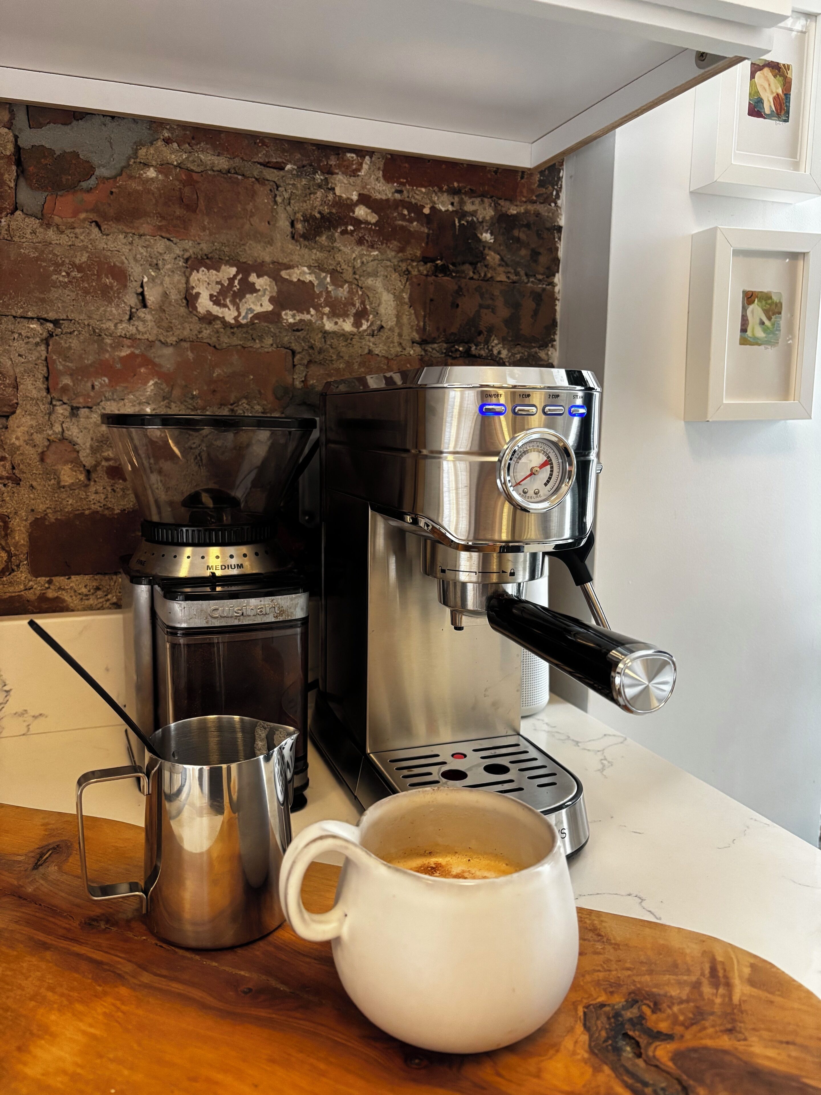 A coffee machine with a cup of coffee on a wooden table, a metal pitcher, and a brick wall in the background.