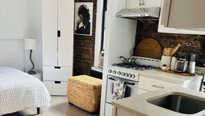 Een compacte slaapkamer met een bed, nachtkastje en lamp. Grenzend daar aan bevindt zich een kitchenette met een kookplaat, spoelbak en bergkasten. In het woongedeelte is een rieten bankje en een kamerkleed met een patroon zichtbaar.