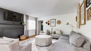 Ein Wohnzimmer mit grauem Sofa, einem gemusterten Ottomanen und einem flachbildfernseher an der Wand.