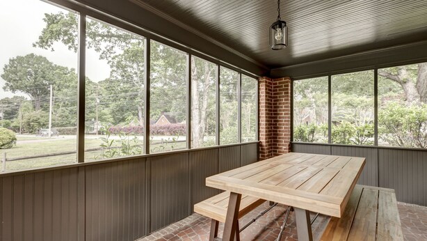 Eine überdachte Terrasse mit Holztisch und Bänken, große Fenster mit Blick auf einen Garten und Bäume.