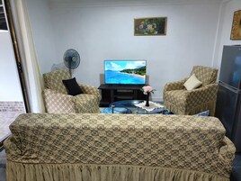 A living room with patterned sofas, a flat-screen TV, a fan, and a vase with flowers.