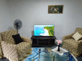 A living room with patterned sofas, a flat-screen TV displaying a beach scene, a fan, and a glass coffee table with a vase of flowers.