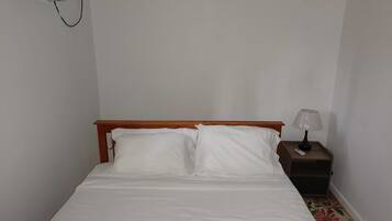 Una cama individual con ropa de cama blanca, cabecera de madera, mesita de noche con lámpara y toma de corriente en la pared.