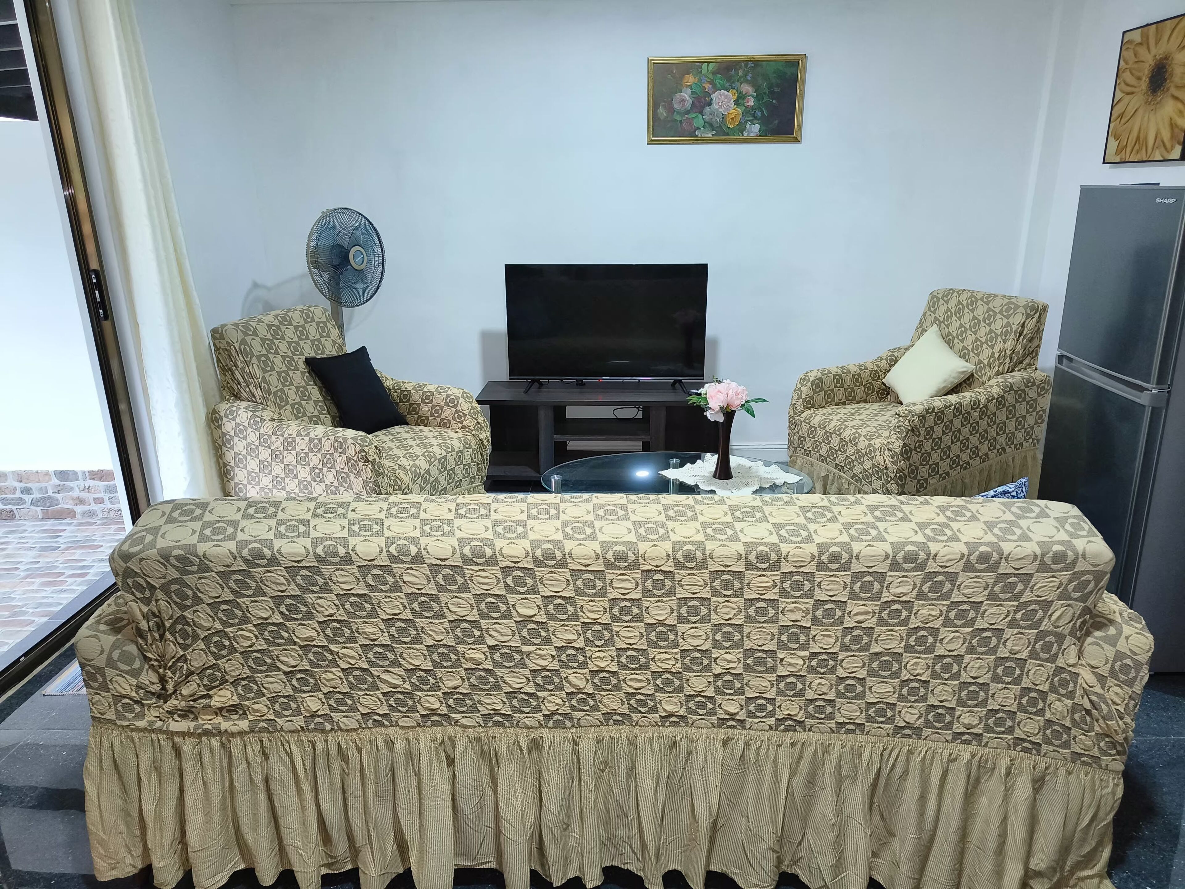 Un salon avec des canapés à motifs, un téléviseur à écran plat, un ventilateur et un réfrigérateur.