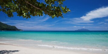 A beach with clear turquoise water, white sand, and a distant mountain.