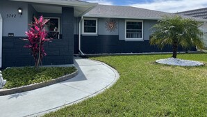 A residential house with a grey and dark grey exterior, a curved driveway, and a small garden with a palm tree and a pink flowering plant. - Sunset Serenity Retreat: Coastal Elegance with Poolside Bliss (New Port Richey)