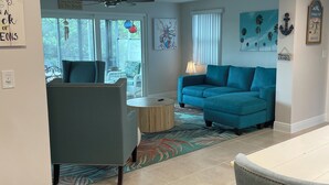 A living room with a blue sofa, a round coffee table, and a dining area with a white table and chairs. There are decorative wall art pieces and a ceiling fan. - Sunset Serenity Retreat: Coastal Elegance with Poolside Bliss (New Port Richey)