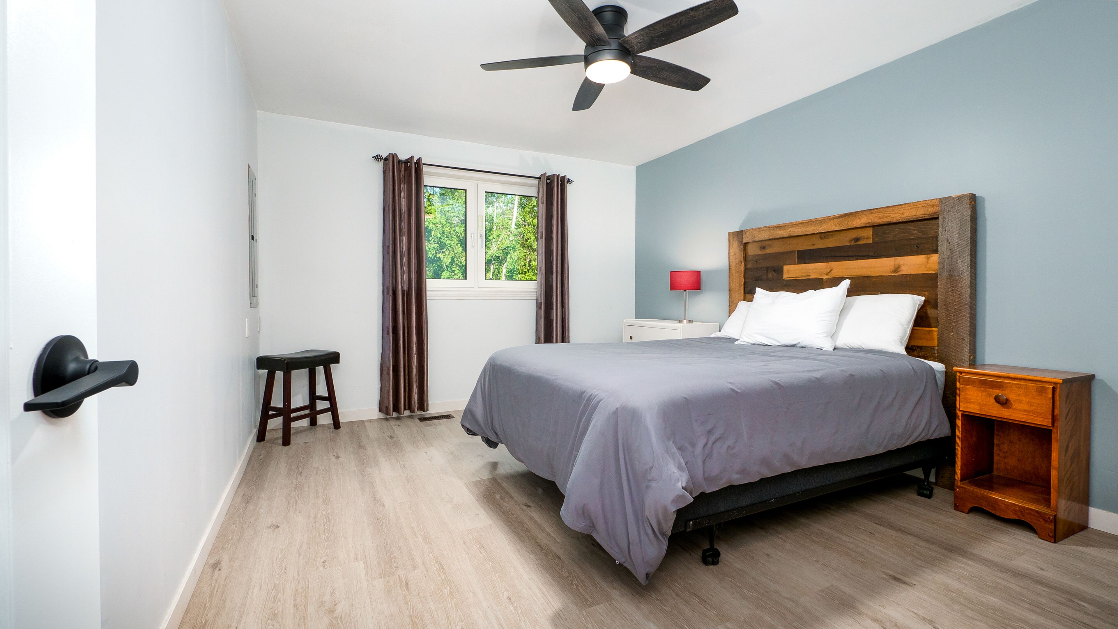 Une chambre à coucher comprenant un lit, une table de chevet, une tête de lit en bois, un ventilateur de plafond, une fenêtre avec des rideaux et une porte.