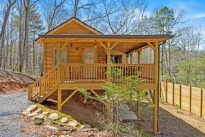 A wooden cabin with a porch, stone foundation, and a view of the surrounding forest.