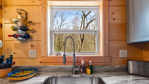 Una cocina con un lavamanos de acero inoxidable, armarios de madera, un ventanal con vistas a árboles y una estantería para vinos con botellas.