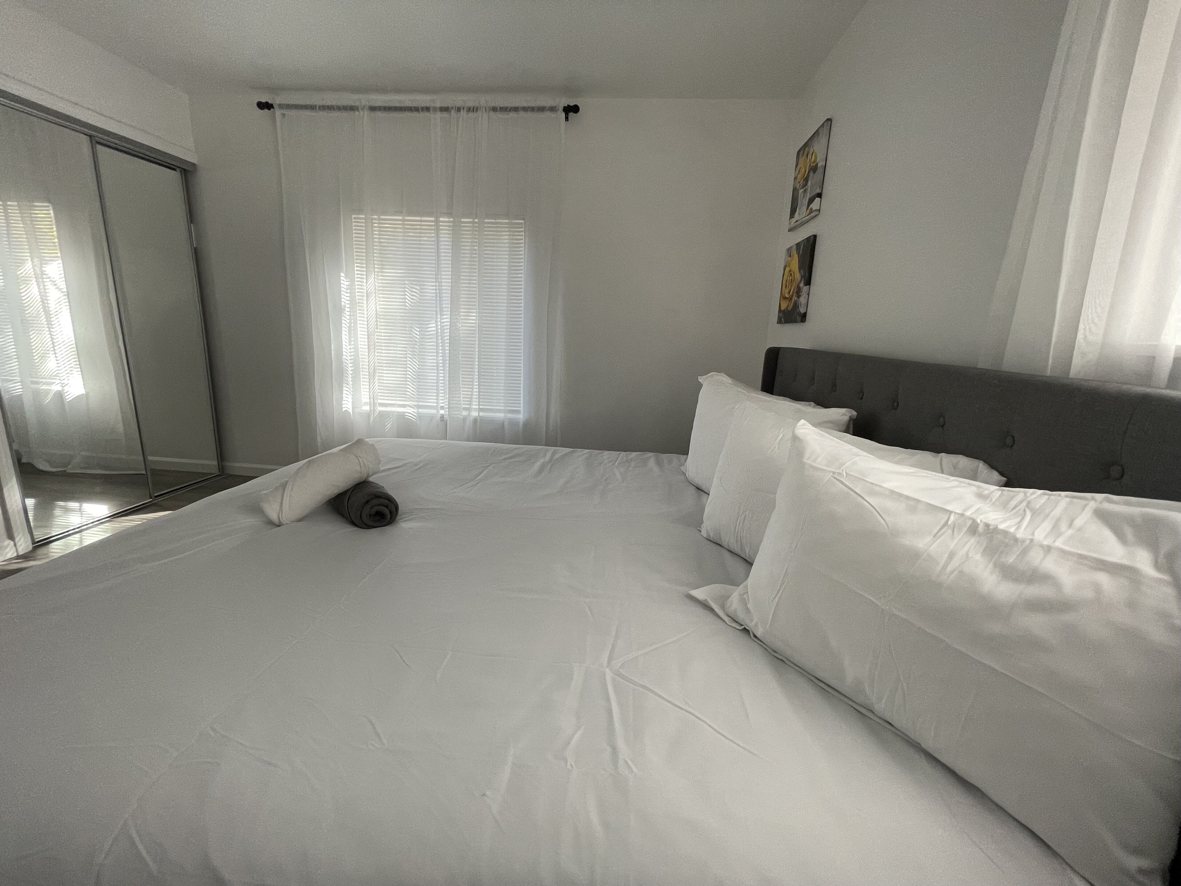 Cama bem arrumada com lençóis e almofadas brancas, cabeceira cinza e janela com persianas.