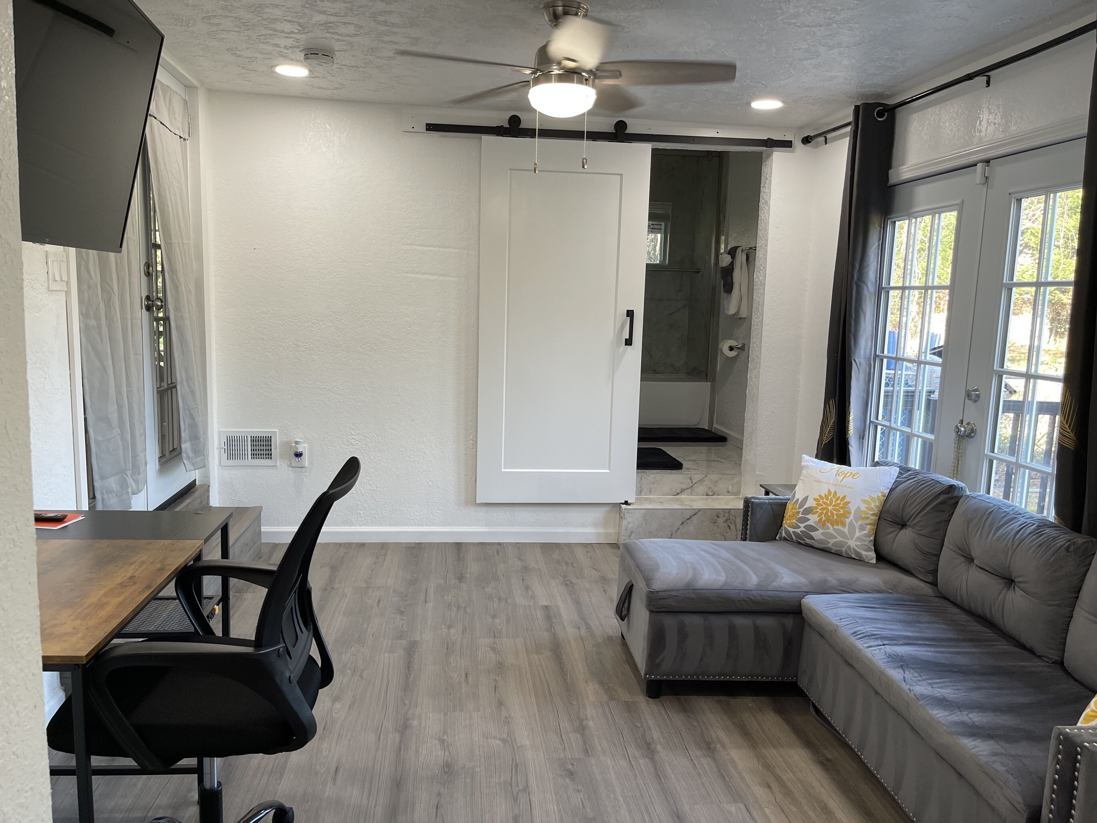 A modern living space with a wooden dining table, black office chair, grey sectional sofa, and a ceiling fan.