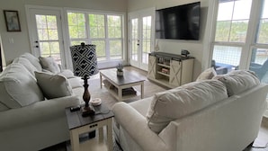 A spacious living room with two sofas, a coffee table, and a TV. Large windows provide natural light and a view of greenery outside.