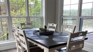 A dining area with a dark wooden table and white chairs, large windows with a view of trees, and a ceiling light fixture.