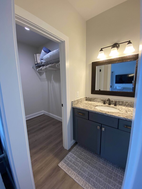Un baño moderno con un espejo grande, un lavabo y tocador. Hay un vestidor con una barra para colgar ropa y una puerta que da a un dormitorio.