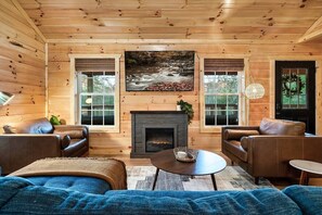 Ein gemütliches Blockhaus-Interieur mit Kamin, Ledersofas und einem großen Fenster mit Blick auf einen Wald.