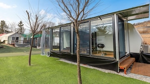 Una casa móvil moderna con paredes de vidrio, un área exterior cubierta, una parrilla de barbacoa y una terraza de madera.