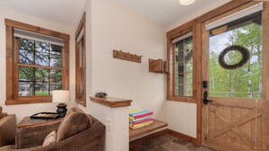 A cozy living room with a brown sofa, a lamp, and a window with a view of trees. There's a wooden door with a wreath and a shelf with books and a tray. - Remodeled Granby Ranch Condo - 20 miles to RMNP w/ Hot Tub & Firepit (Granby)