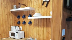 Une cuisine compacte aux murs en bois, équipée d’un four à micro-ondes, d’un grille-pain, d’un évier et d’étagères où sont rangées des tasses et des assiettes.