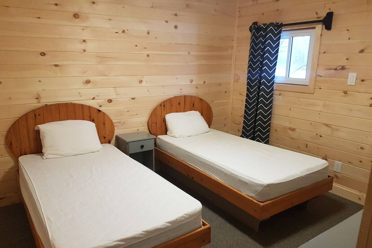 Een blokhut met twee houten bedden, een klein nachtkastje en een raam met gordijn.