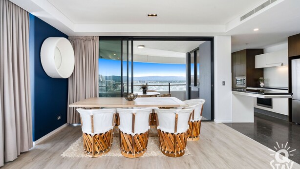 A modern dining area with a wooden table and white chairs, a large window with a city view, and a kitchen in the adjacent room. - The Wave - Q Stay (Broadbeach)