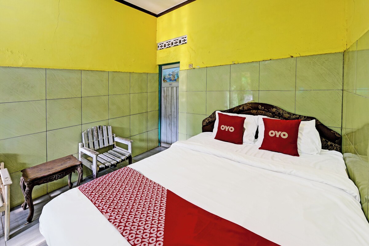 Habitación de hotel con una cama grande, dos almohadas rojas con el logo 'OYO' y una silla de madera, además de paredes con azulejos verdes.