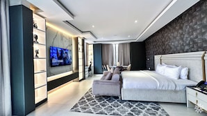 A modern bedroom with a large bed, a TV mounted on the wall, and a seating area with a sofa and chairs.