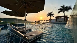 A pool area with lounge chairs and umbrellas, a water fountain, palm trees, and a sunset in the background.