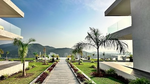A modern building with a central pathway flanked by greenery and palm trees, leading to a scenic view of mountains.