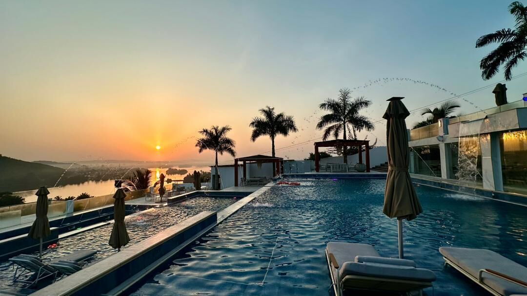 A pool area with lounge chairs and umbrellas, overlooking a body of water and palm trees during sunset.