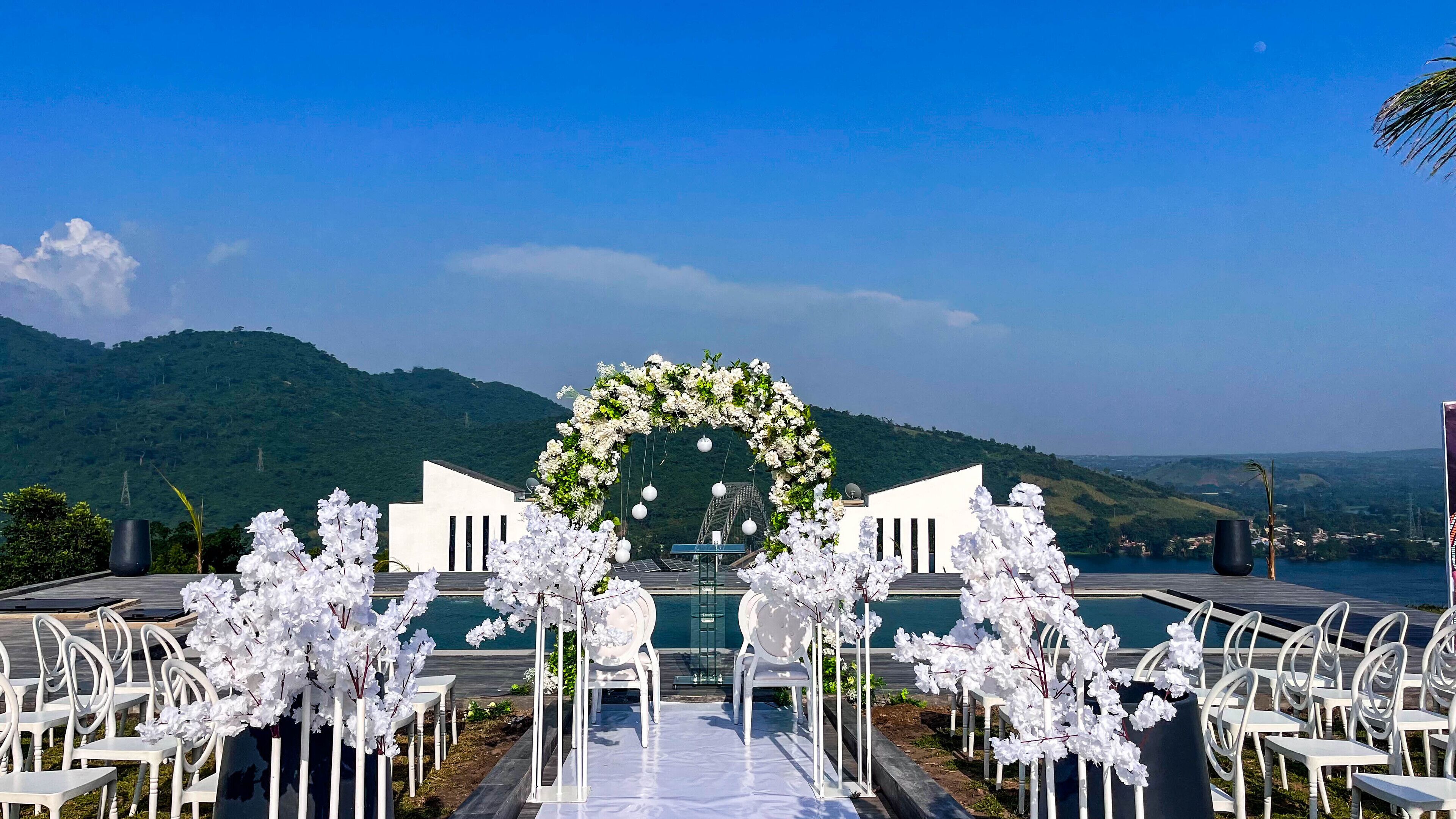 Une cérémonie de mariage en plein air, avec des arches et des chaises décorées de fleurs blanches, offrant une vue sur un plan d’eau et des collines.