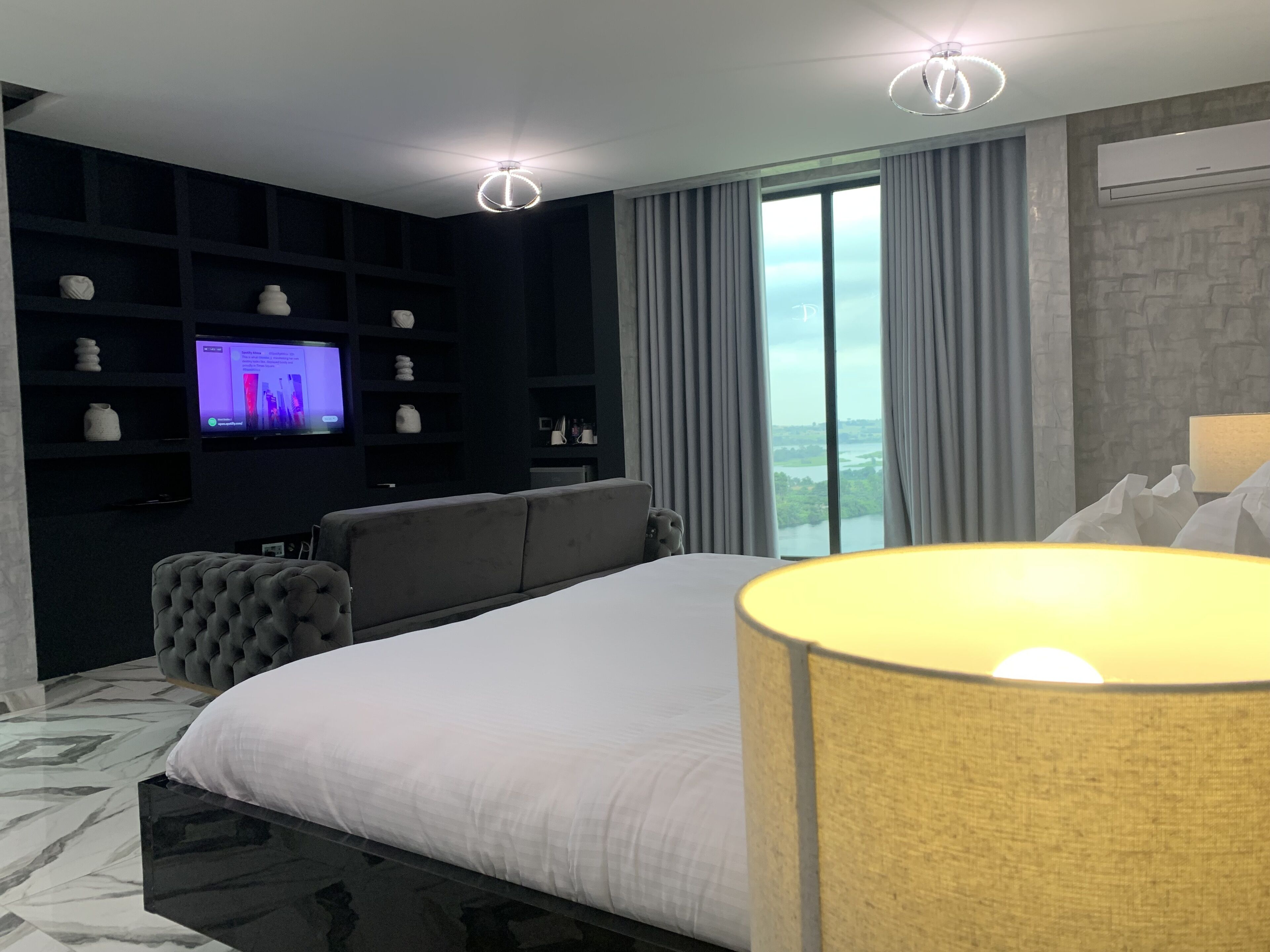 A modern hotel room with a large bed, a television on a dark wall, a sofa, and a view of a cityscape through large windows.