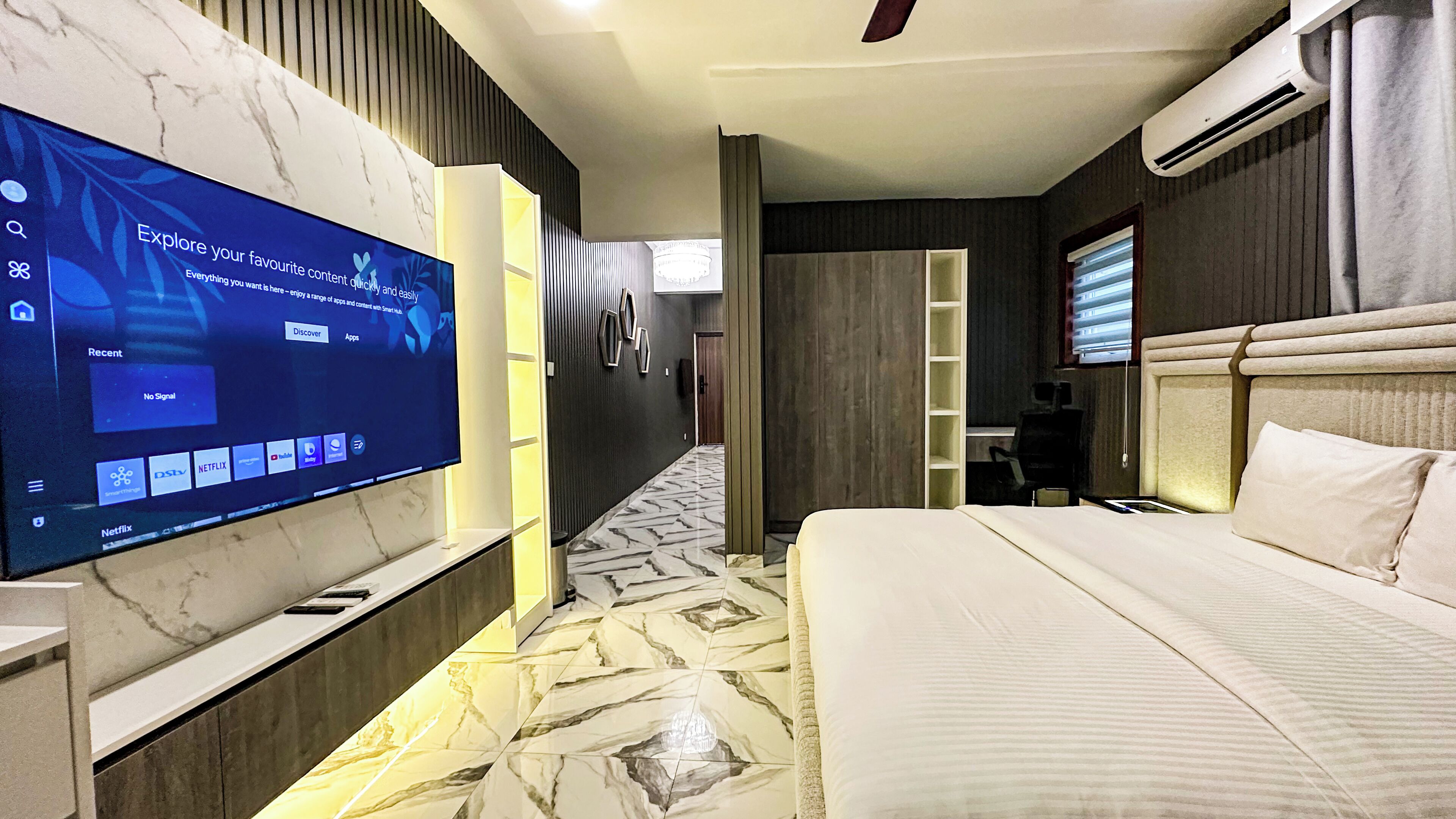 A modern hotel room with a large flat-screen TV mounted on the wall, a bed with a headboard, a desk with a chair, and a hallway with a patterned floor.