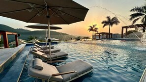 A pool area with lounge chairs and umbrellas, surrounded by palm trees and a sunset view. - BridgeView Resort (Akosombo)