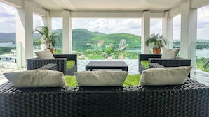 A covered outdoor seating area with wicker furniture and potted plants, overlooking a scenic landscape with hills and a bridge.