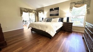Un espacioso dormitorio con pisos de madera, una cama grande, una mesita de noche, un aparador y una ventana con cortinas.