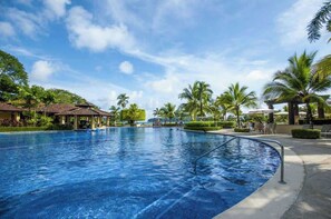 A large swimming pool with a clear blue sky, palm trees, and a covered area with chairs.