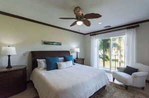 A bedroom with a large bed, two bedside tables with lamps, a ceiling fan, a window with curtains, and a view of greenery outside.