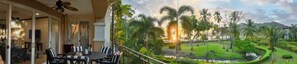 A balcony with a dining table and chairs, a ceiling fan, and a view of a landscaped garden with palm trees and a pond.