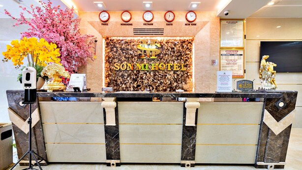 A hotel reception desk with a modern design, featuring a marble counter, a decorative floral arrangement, and a digital clock display. - S79 Son Mi 839 Le Hong Phong Street (Ho Chi Minh City)