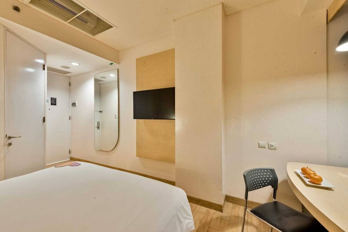A hotel room with a bed, a chair, a small table with a plate of food, a TV mounted on the wall, and a door leading to another room.