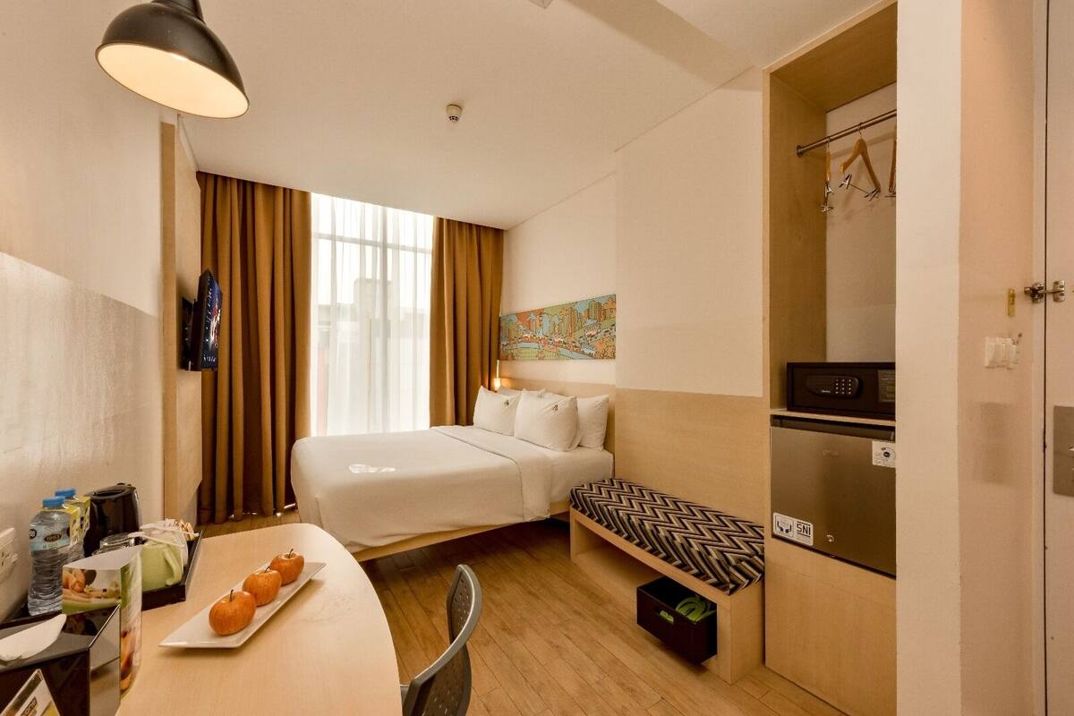 A compact hotel room with a bed, a small desk with a chair, a dining table with apples, a refrigerator, and a microwave.