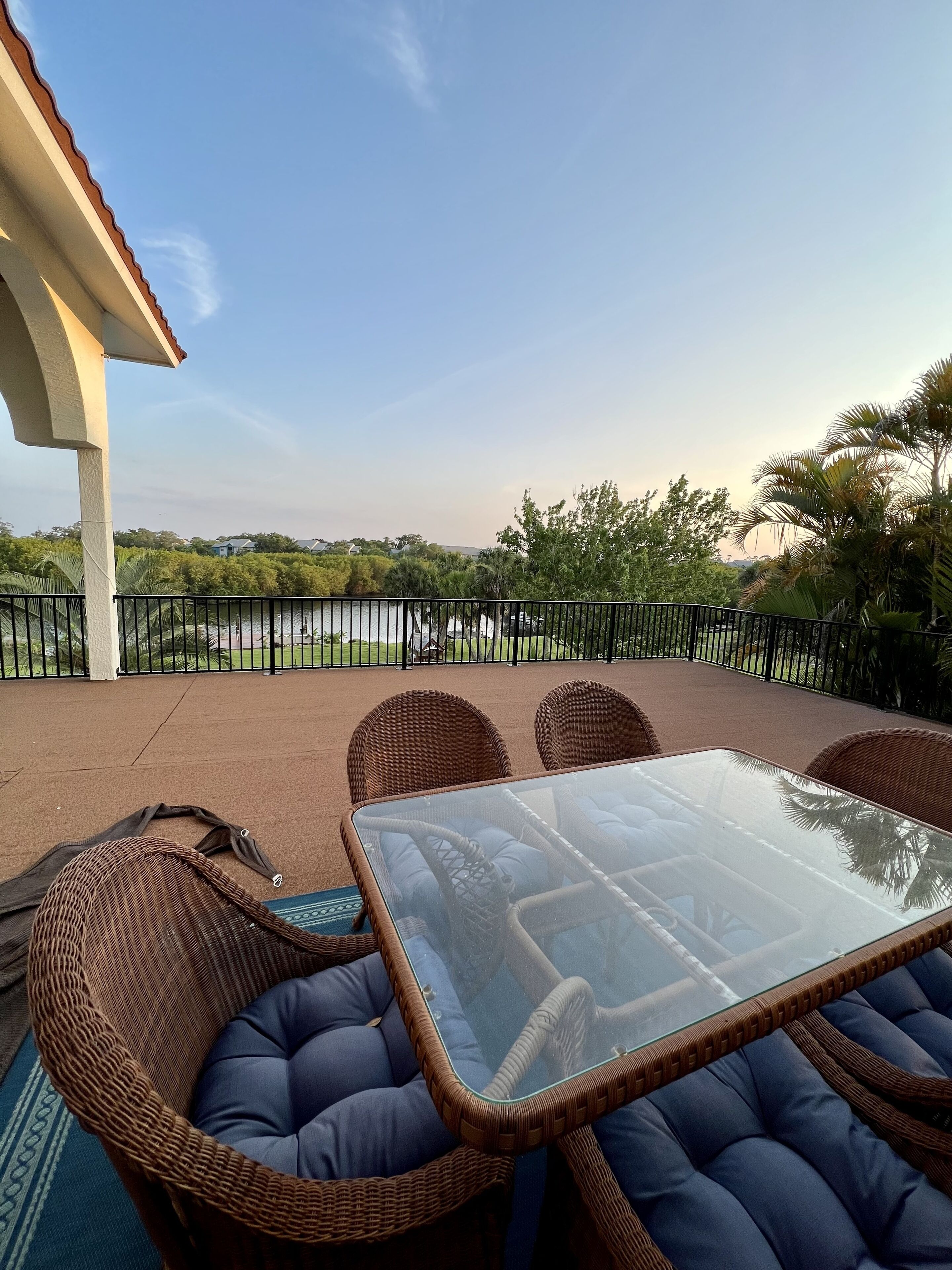 Eine Terrasse mit Korbmöbeln und einem Glastisch, mit Blick auf eine angelegte Grünfläche mit Bäumen und einem Horizont.