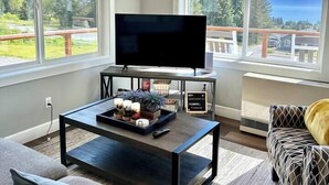Een moderne woonkamer met een flatscreen televisie op een bijzettafeltje, een bank en een gestreept fauteuil. Grote ramen bieden uitzicht op een bos en bergen.