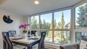 A dining area with a table set for four, large windows, and a view of trees and a house. - Le Chamois 211 - Ski in Ski out, Common Pool, Hot Tub & Gym (Whistler)