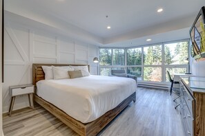 A modern bedroom with a large bed, a desk, and a view of trees through the windows.