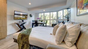 A modern bedroom with a large bed, a desk, and a view of the outdoors through large windows. - Le Chamois 211 - Ski in Ski out, Common Pool, Hot Tub & Gym (Whistler)