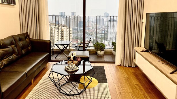 Un salon moderne avec un canapé, une table basse et un téléviseur. Il y a un balcon avec vue sur la ville.