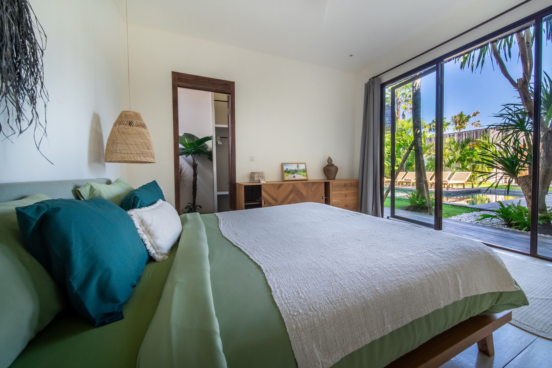 A bedroom with a bed, a dresser, and a large window with a view of a garden.